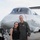 Two people pose together in front of a military or government aircraft on a tarmac. The taller individual is wearing an olive green flight suit with patches and wings insignia, consistent with a military aviator. The other person is dressed in civilian clothing — a black top and jeans with sunglasses.