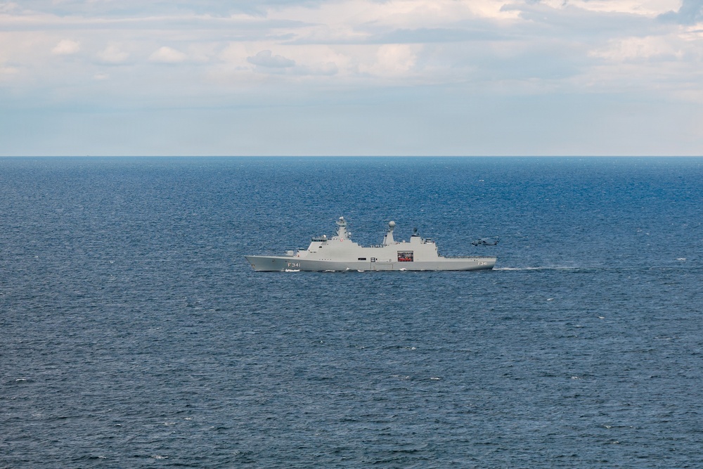 A gray naval frigate bearing the hull number F341 is underway on open blue ocean waters. The vessel appears to be a modern warship with visible radar and communication masts, and what appears to be a helicopter in close proximity to the ship's stern. The sky is partly cloudy with calm sea conditions.
