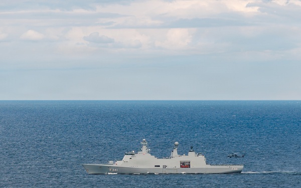 A gray naval frigate bearing the hull number F341 is underway on open blue ocean waters. The vessel appears to be a modern warship with visible radar and communication masts, and what appears to be a helicopter in close proximity to the ship's stern. The sky is partly cloudy with calm sea conditions.