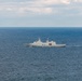 A gray naval frigate bearing the hull number F341 is underway on open blue ocean waters. The vessel appears to be a modern warship with visible radar and communication masts, and what appears to be a helicopter in close proximity to the ship's stern. The sky is partly cloudy with calm sea conditions.