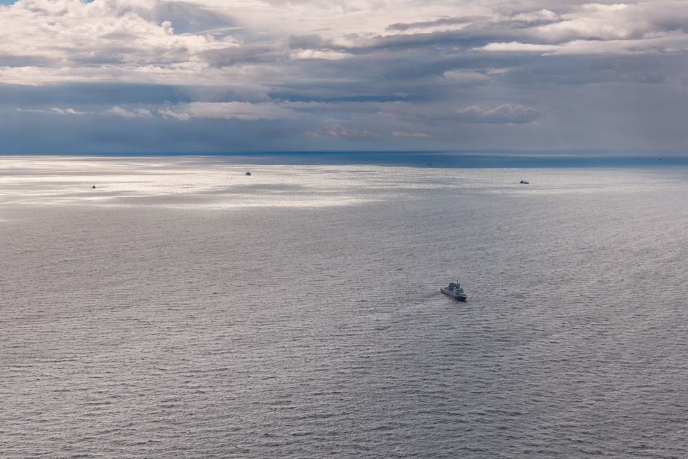 An aerial or elevated view of a large open body of water under a partly cloudy sky with dramatic cloud formations. A medium-sized vessel is visible in the foreground-middle area, with at least three smaller vessels visible near the horizon. The water surface is calm with gentle ripples, reflecting diffused light through breaks in the clouds.