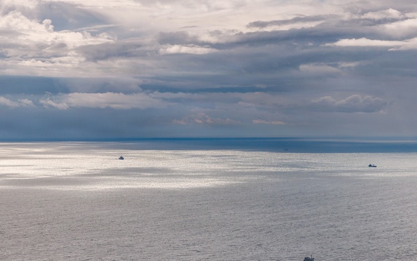 An aerial or elevated view of a large open body of water under a partly cloudy sky with dramatic cloud formations. A medium-sized vessel is visible in the foreground-middle area, with at least three smaller vessels visible near the horizon. The water surface is calm with gentle ripples, reflecting diffused light through breaks in the clouds.