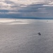 An aerial or elevated view of a large open body of water under a partly cloudy sky with dramatic cloud formations. A medium-sized vessel is visible in the foreground-middle area, with at least three smaller vessels visible near the horizon. The water surface is calm with gentle ripples, reflecting diffused light through breaks in the clouds.