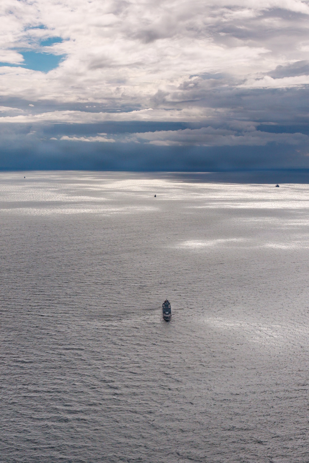 An aerial or elevated view of a large open body of water under a dramatic, partially cloudy sky with patches of blue and dark storm clouds. A larger vessel is visible in the foreground-middle area, with at least two smaller vessels visible further in the distance near the horizon. Silver light reflects off the water's surface in scattered patches, creating a striking atmospheric effect.