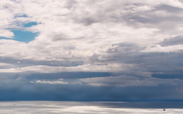 An aerial or elevated view of a large open body of water under a dramatic, partially cloudy sky with patches of blue and dark storm clouds. A larger vessel is visible in the foreground-middle area, with at least two smaller vessels visible further in the distance near the horizon. Silver light reflects off the water's surface in scattered patches, creating a striking atmospheric effect.