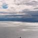 An aerial or elevated view of a large open body of water under a dramatic, partially cloudy sky with patches of blue and dark storm clouds. A larger vessel is visible in the foreground-middle area, with at least two smaller vessels visible further in the distance near the horizon. Silver light reflects off the water's surface in scattered patches, creating a striking atmospheric effect.
