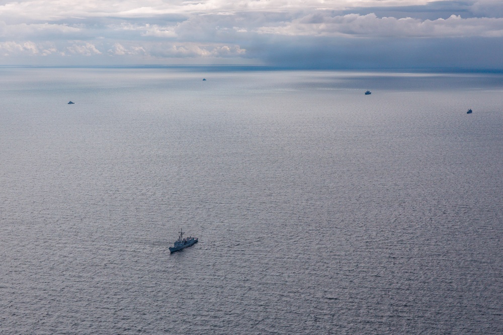 An aerial photograph shows multiple naval or military vessels spread across a calm, open sea under an overcast sky with dramatic cloud formations. One larger warship is prominently visible in the foreground-center, while four additional smaller vessels are distributed across the middle and far distance. The muted blue-grey tones of the water and sky suggest overcast lighting conditions, possibly at dusk or under heavy cloud cover.