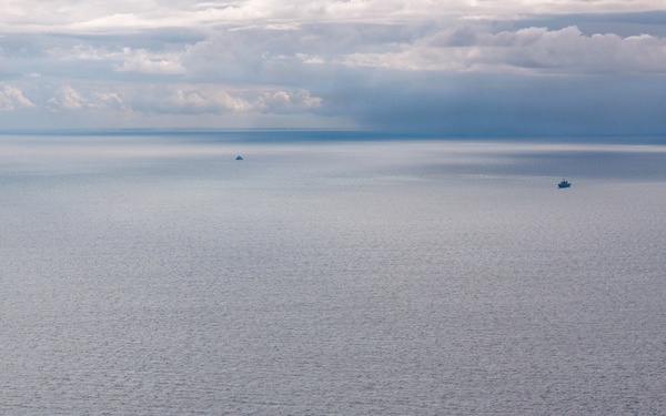 An aerial photograph shows multiple naval or military vessels spread across a calm, open sea under an overcast sky with dramatic cloud formations. One larger warship is prominently visible in the foreground-center, while four additional smaller vessels are distributed across the middle and far distance. The muted blue-grey tones of the water and sky suggest overcast lighting conditions, possibly at dusk or under heavy cloud cover.