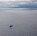An aerial photograph shows multiple naval or military vessels spread across a calm, open sea under an overcast sky with dramatic cloud formations. One larger warship is prominently visible in the foreground-center, while four additional smaller vessels are distributed across the middle and far distance. The muted blue-grey tones of the water and sky suggest overcast lighting conditions, possibly at dusk or under heavy cloud cover.