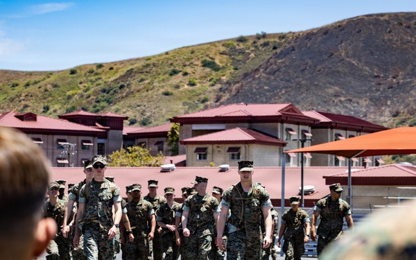 A group of military personnel in woodland camouflage uniforms are marching in formation across a paved area at what appears to be a military installation. The scene is observed from behind by other service members in the foreground. The setting includes low-rise buildings with red roofs and a hillside in the background, consistent with a U.S. Marine Corps base.