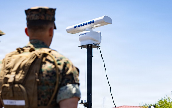 Military personnel in camouflage uniforms are gathered around a Furuno NXT radar unit mounted on a tripod stand during what appears to be a field demonstration or equipment briefing. A solar panel is visible at the base of the radar unit, suggesting a portable, self-powered setup. The scene takes place outdoors in a military or training environment with vehicles and structures visible in the background.