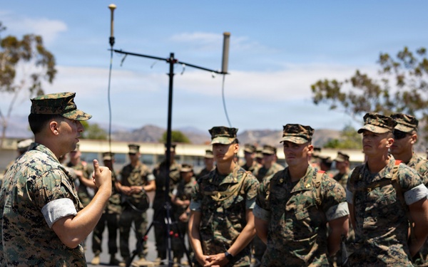 A military instructor or senior non-commissioned officer in MARPAT camouflage uniform addresses a formation of Marines in an outdoor setting. The speaker appears to be conducting a briefing or instruction session, with personnel standing in a semi-circular formation listening attentively. The setting appears to be a military installation, likely a base in Southern California based on the terrain and vegetation.
