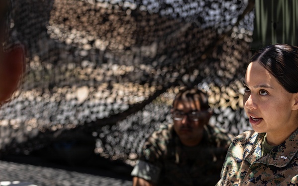A female U.S. Marine in MARPAT woodland digital camouflage uniform is speaking or briefing others under a camouflage netting shade structure. At least two other military personnel are visible in the background, one seated and wearing sunglasses. The setting appears to be an outdoor field environment, likely during a training exercise or operational briefing.