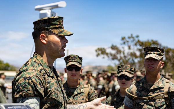 A U.S. Marine in digital camouflage (MARPAT) uniform is conducting a briefing or demonstration for a group of fellow Marines in an outdoor setting. He is gesturing toward equipment that includes a solar panel and what appears to be a weather station or sensor system mounted on a tripod. The setting appears to be a military installation, likely in a Southern California environment based on the terrain and vegetation.