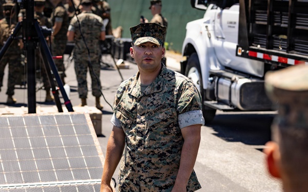 A U.S. Marine in MARPAT camouflage uniform appears to be conducting a briefing or demonstration involving solar panels laid out on the ground. The setting is an outdoor military environment with multiple uniformed personnel gathered in the background. A laptop and portable power equipment are visible nearby, suggesting a field energy systems training or demonstration.