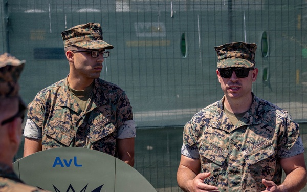 A group of U.S. Marines in MARPAT digital camouflage uniforms are gathered outdoors for what appears to be a briefing or training session. One Marine, wearing sunglasses with a name tape reading 'O'BRIEN' and 'U.S. MARINES,' is speaking to the group while gesturing with his hands. A satellite dish or antenna dish marked 'AvL' and 'NO LIFT' is visible being held by one of the Marines.