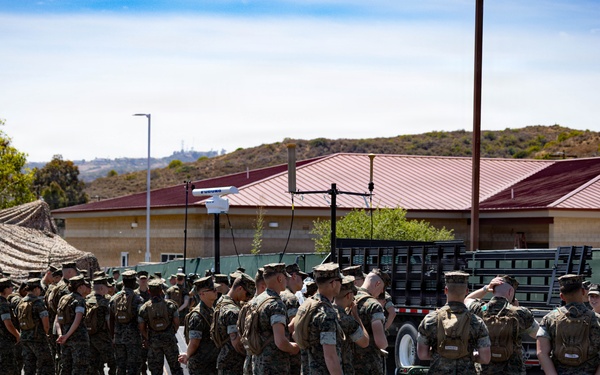 A large group of military personnel in camouflage uniforms are assembled in formation on a paved area at what appears to be a U.S. Marine Corps base. The personnel are gathered near military vehicles and equipment, including a camouflage net structure and a flatbed truck. The setting includes base buildings with red roofs and hilly terrain in the background, consistent with a Southern California military installation.