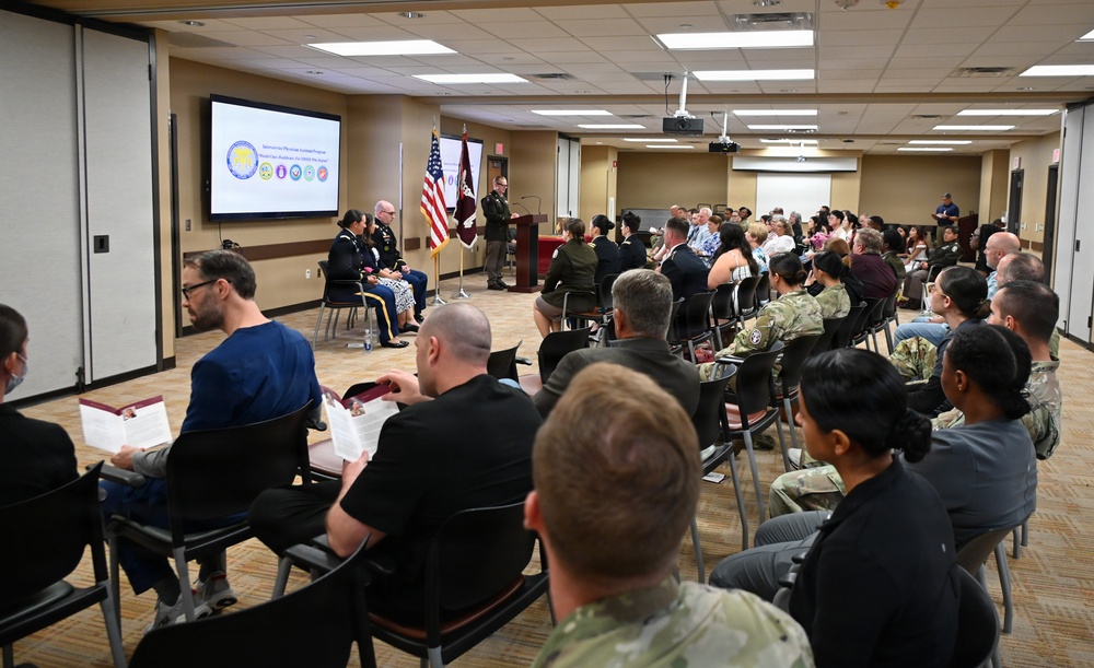 A formal military briefing or ceremony is taking place in a conference room, with a speaker at a podium addressing a mixed audience of military personnel in uniform and civilians. A presentation slide is displayed on a large screen showing what appears to be a military healthcare or exchange program logo. Two military officers are seated on the stage area alongside the speaker.