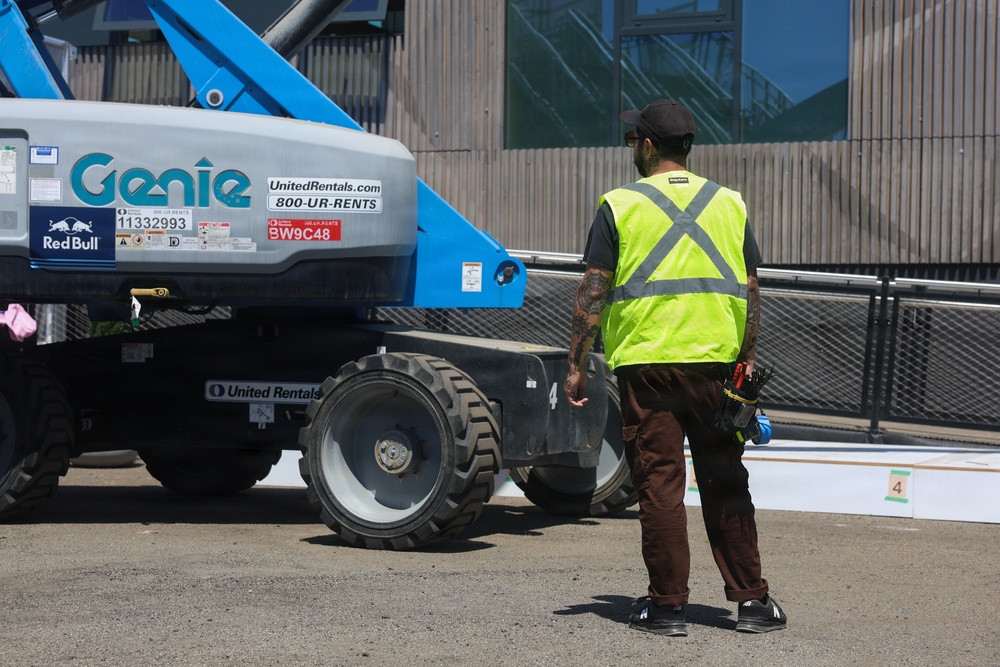 A construction worker wearing a high-visibility safety vest stands near a Genie boom lift rented from United Rentals at what appears to be a construction or event setup site. The worker, who has visible tattoos on their arms, is facing the equipment and appears to be monitoring or directing its operation. The setting is an outdoor paved area adjacent to a modern building with glass and wood facade elements.