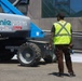 A construction worker wearing a high-visibility safety vest stands near a Genie boom lift rented from United Rentals at what appears to be a construction or event setup site. The worker, who has visible tattoos on their arms, is facing the equipment and appears to be monitoring or directing its operation. The setting is an outdoor paved area adjacent to a modern building with glass and wood facade elements.