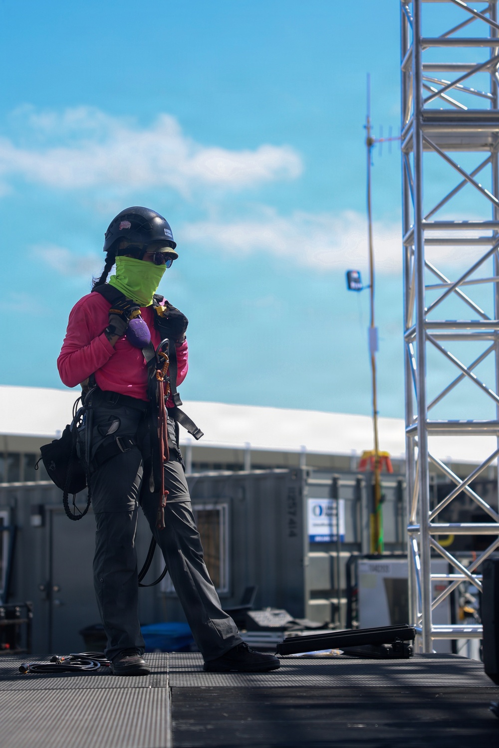 A worker in full personal protective equipment stands on an elevated industrial platform, adjusting their fall-arrest harness and rigging gear. The individual is equipped for high-elevation or confined-space work, wearing a hard hat, face gaiter, safety glasses, and a full-body harness with carabiners. The background reveals an industrial or construction site with metal scaffolding/truss structures, equipment units, and a communications antenna.