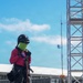 A worker in full personal protective equipment stands on an elevated industrial platform, adjusting their fall-arrest harness and rigging gear. The individual is equipped for high-elevation or confined-space work, wearing a hard hat, face gaiter, safety glasses, and a full-body harness with carabiners. The background reveals an industrial or construction site with metal scaffolding/truss structures, equipment units, and a communications antenna.