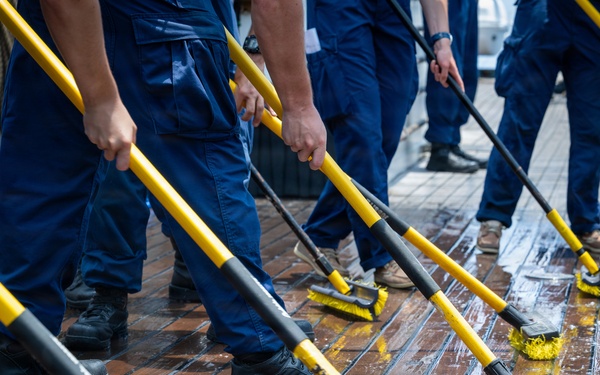 A group of uniformed personnel in navy blue coveralls are scrubbing a wet wooden deck in a coordinated, line-formation manner. They are using identical yellow-handled deck brushes, suggesting a military or maritime training exercise. The teak or hardwood deck appears to be on a ship or vessel, with soapy water and cleaning residue visible.