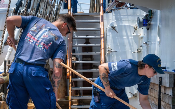 Two personnel in blue uniforms are performing maintenance work on the wooden deck of what appears to be a tall ship or sailing vessel. They are using long-handled tools, likely holystones or scrubbing brushes, alongside a bucket of cleaning or caulking material. The cap of one individual bears 'U.S. Coast Guard Eagle' markings, identifying this as the USCGC Eagle training vessel.