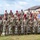 A large group of U.S. Army soldiers in camouflage uniforms and distinctive maroon berets are assembled outdoors in a formation, appearing to be in a moment of prayer or reflection with heads bowed. They are standing in front of bleachers near a military installation building that displays crossed-axes insignia. The setting appears to be a formal military ceremony or memorial event.