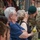 A U.S. Army soldier wearing a green beret and multicam uniform is leaning down to kiss or greet a civilian woman who is holding yellow roses. The soldier's name tape reads 'HEISLER' and he wears a Special Forces-style patch on his shoulder. This appears to be a military ceremony or homecoming event with family members and other soldiers visible in the background.