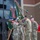 A military ceremony is taking place in front of a modern brick building, involving several soldiers in camouflage uniforms and green berets. Multiple military flags and guidons are prominently displayed, including a US flag and green unit colors adorned with campaign streamers. This appears to be a change of command or activation/inactivation ceremony typical of US Army Special Forces units.