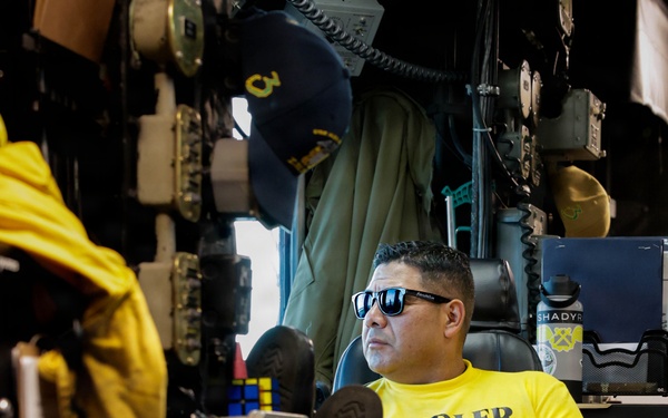 A man wearing a bright yellow long-sleeve shirt labeled 'HANDLER' and reflective sunglasses sits in a chair in what appears to be a naval vessel workspace or control room. A yellow flight deck handler's jersey with 'HANDLER' markings hangs in the foreground. The environment is cluttered with equipment, cables, a calendar, a water bottle, and various naval gear.