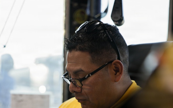 A middle-aged man wearing a yellow long-sleeve shirt and black-framed glasses is seated in what appears to be an enclosed vehicle or equipment cab. He is looking downward and writing or reviewing documents with a pen in hand. The background shows an outdoor environment visible through a window, with various equipment and controls visible to the left.