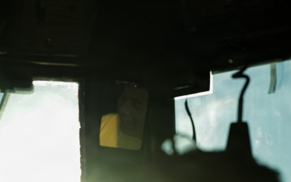 The image is taken from inside a vehicle or enclosed cab, looking toward a bright exterior light source causing significant lens flare. A person wearing a yellow garment is partially visible in what appears to be a rearview or side mirror. The interior is heavily shadowed, with silhouetted forms dominating the foreground.
