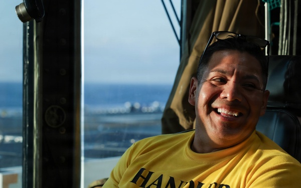 A smiling man wearing a yellow long-sleeve shirt labeled 'HANDLER' is seated in what appears to be an elevated control station or tower with a view of the ocean and a flight deck or airfield in the background. He is holding a radio or communication device and has sunglasses resting on top of his head. The setting suggests a military or aviation operations environment.