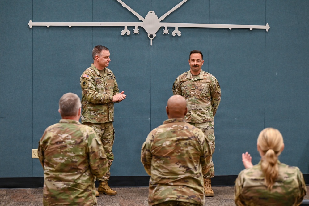 A military ceremony or recognition event is taking place indoors, with two service members standing at the front facing an audience of fellow personnel. A large wall-mounted silhouette of what appears to be an MQ-9 Reaper drone serves as a backdrop. The audience members are applauding, suggesting an award presentation or commendation ceremony.