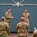 A military ceremony or recognition event is taking place indoors, with two service members standing at the front facing an audience of fellow personnel. A large wall-mounted silhouette of what appears to be an MQ-9 Reaper drone serves as a backdrop. The audience members are applauding, suggesting an award presentation or commendation ceremony.