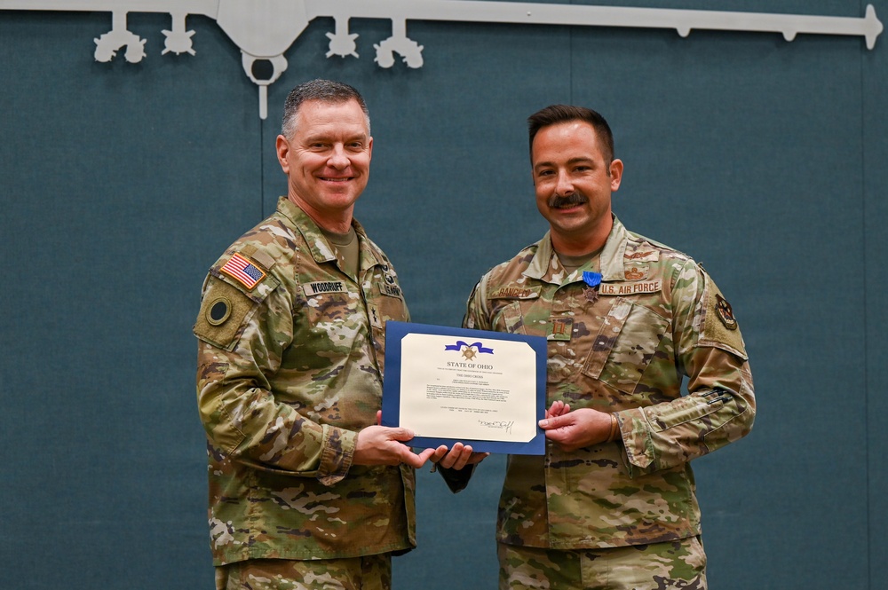 Two military personnel in OCP (Operational Camouflage Pattern) uniforms are posing together holding a certificate from the State of Ohio. The individual on the right is wearing a medal on his chest, suggesting this is an awards ceremony. The backdrop features a silhouette outline of a military aircraft on a teal/green panel.