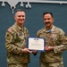Two military personnel in OCP (Operational Camouflage Pattern) uniforms are posing together holding a certificate from the State of Ohio. The individual on the right is wearing a medal on his chest, suggesting this is an awards ceremony. The backdrop features a silhouette outline of a military aircraft on a teal/green panel.