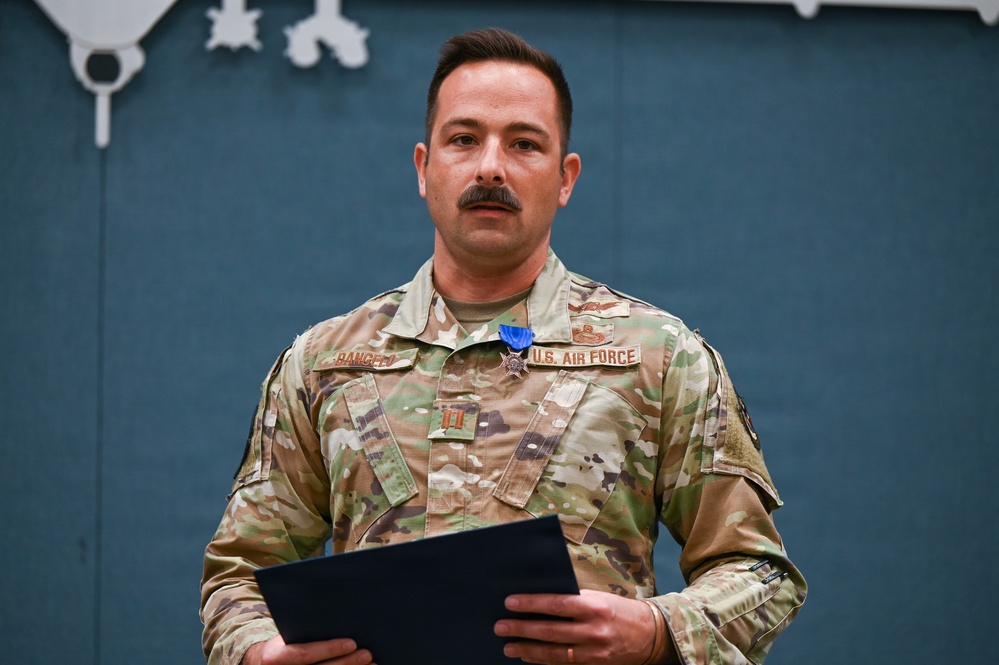 A U.S. Air Force service member in OCP (Operational Camouflage Pattern) uniform is standing at attention during what appears to be a formal military ceremony. He is wearing a medal with a blue ribbon, consistent with a decoration presentation, and holds a black folder or certificate. His name tape reads 'RANGEL' and he wears captain's rank insignia.