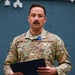 A U.S. Air Force service member in OCP (Operational Camouflage Pattern) uniform is standing at attention during what appears to be a formal military ceremony. He is wearing a medal with a blue ribbon, consistent with a decoration presentation, and holds a black folder or certificate. His name tape reads 'RANGEL' and he wears captain's rank insignia.