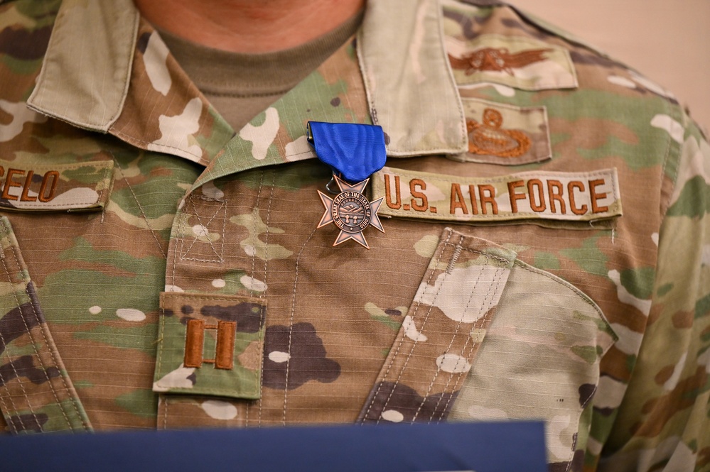 A close-up photograph of a U.S. Air Force service member's uniform chest area, showing an OCP (Operational Camouflage Pattern) uniform with a medal pinned above the name tape. The medal has a blue ribbon and appears to be a cross/star-shaped award, likely a state-level or military commendation medal. Rank insignia visible on the left chest pocket indicates the rank of Captain (O-3).