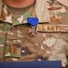 A close-up photograph of a U.S. Air Force service member's uniform chest area, showing an OCP (Operational Camouflage Pattern) uniform with a medal pinned above the name tape. The medal has a blue ribbon and appears to be a cross/star-shaped award, likely a state-level or military commendation medal. Rank insignia visible on the left chest pocket indicates the rank of Captain (O-3).