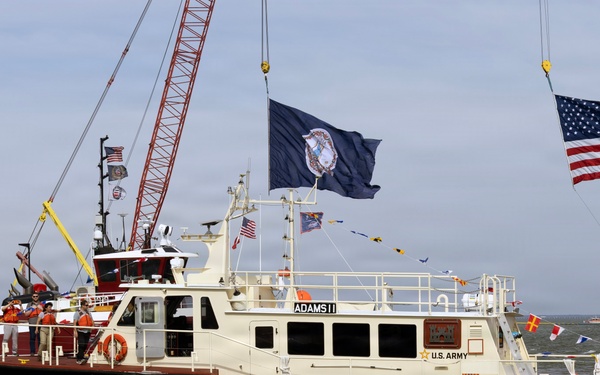 A U.S. Army Corps of Engineers vessel named 'Adams II' is underway on open water, appearing to be part of a commissioning or christening ceremony. The boat is decorated with multiple flags including a large U.S. flag and what appears to be a U.S. Army flag, along with signal pennants. Several crew members in orange safety vests are visible on deck, and industrial cranes are visible in the background.