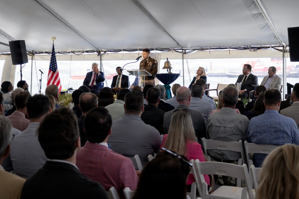 A formal outdoor ceremony is taking place under a large white tent, likely near a waterfront or port facility. A person in military uniform is speaking at a podium equipped with a microphone, while dignitaries and officials are seated on a stage behind them. A large audience seated in white folding chairs faces the speaker.