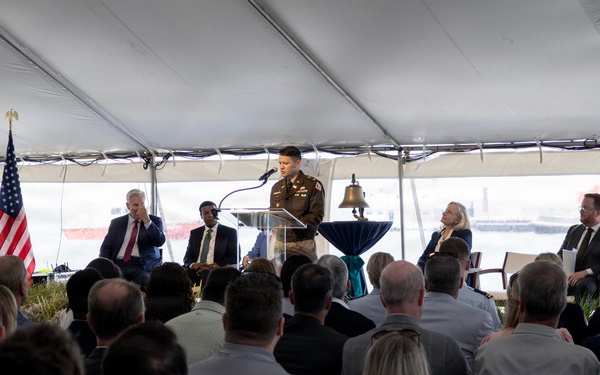 A formal outdoor ceremony is taking place under a large white tent, likely near a waterfront or port facility. A person in military uniform is speaking at a podium equipped with a microphone, while dignitaries and officials are seated on a stage behind them. A large audience seated in white folding chairs faces the speaker.