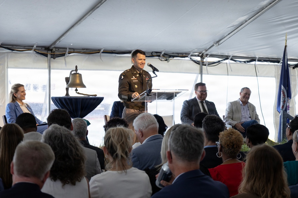 A military officer in U.S. Army dress uniform is speaking at a podium during a formal ceremony held under a large white tent. The event appears to be an official dedication, commemoration, or civic ceremony, evidenced by the presence of a ceremonial bell, a Virginia state flag, and a seated audience of civilians and dignitaries. Several individuals are seated on a dais behind the speaker.