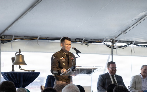 A military officer in U.S. Army dress uniform is speaking at a podium during a formal ceremony held under a large white tent. The event appears to be an official dedication, commemoration, or civic ceremony, evidenced by the presence of a ceremonial bell, a Virginia state flag, and a seated audience of civilians and dignitaries. Several individuals are seated on a dais behind the speaker.