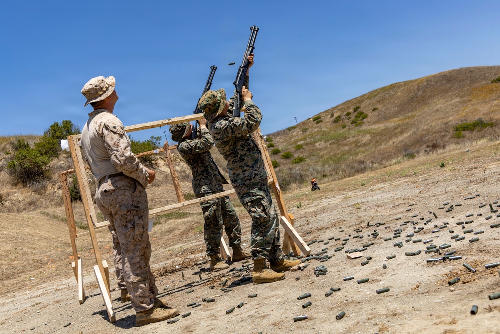 Three military personnel in MARPAT camouflage uniforms are conducting a firearms training exercise at an outdoor shooting range. Two shooters are firing shotguns skyward through a wooden frame structure while a third observes or instructs. Numerous spent shell casings are scattered across the ground, indicating significant prior firing activity.