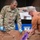 A U.S. Army soldier, identified by name tape as 'MOYA' and wearing Staff Sergeant rank insignia with a 1st Infantry Division patch, works alongside a civilian in an orange shirt to inspect or sort damaged cardboard boxes. The setting appears to be a USPS mail processing or distribution facility, with blue USPS sorting bins and packages visible in the background. The soldier is wearing blue latex gloves and holding a handheld scanner device.